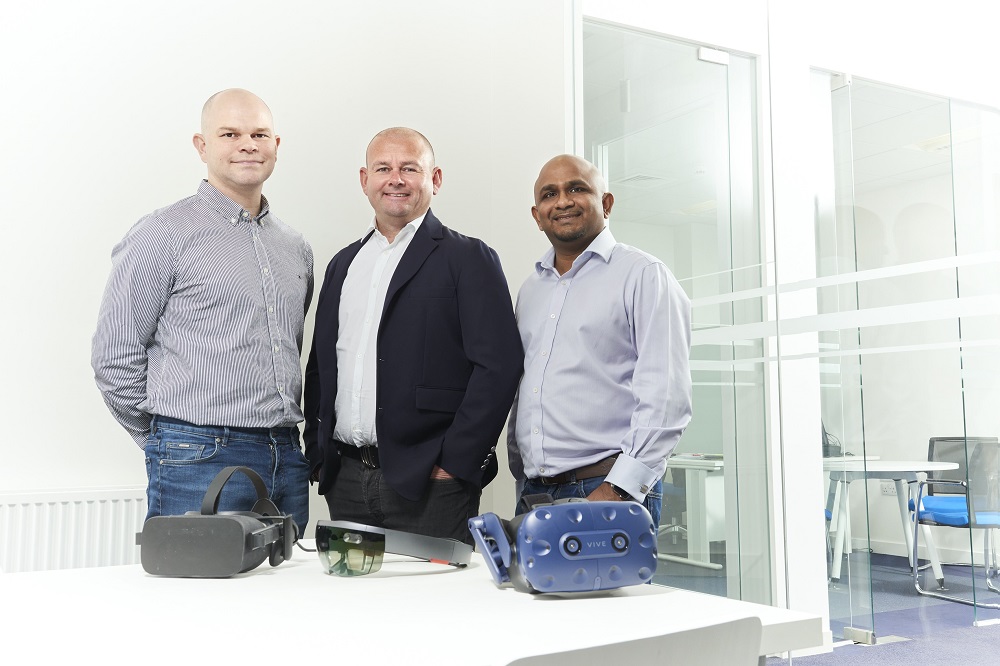 Three men stand in an office beside a table with various virtual reality headsets, including an HTC Vive and other VR devices. The room has a modern design with glass partitions.