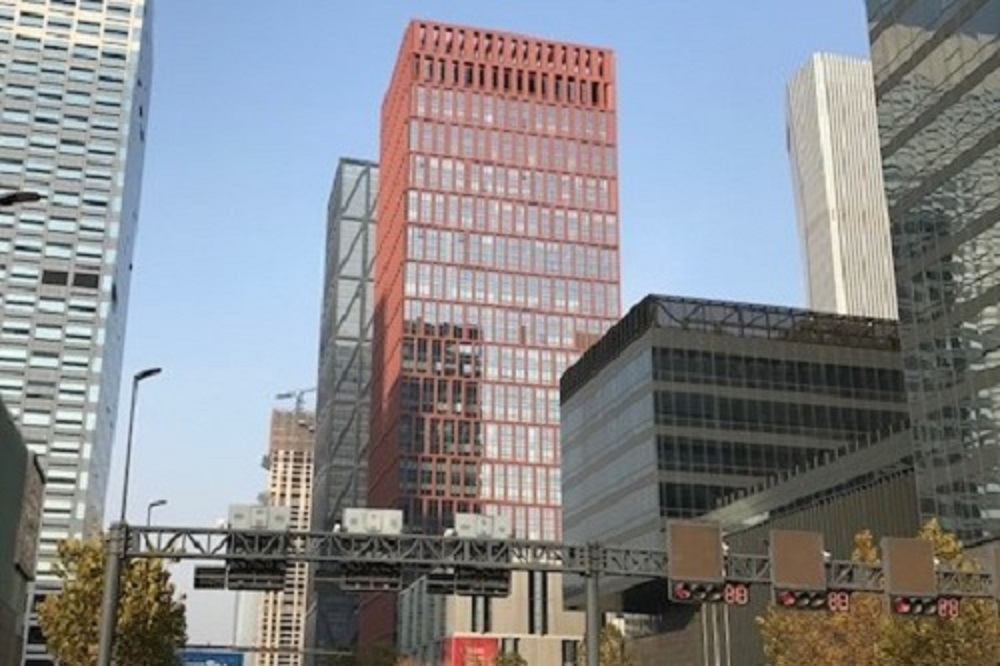 A cityscape featuring a red high-rise building flanked by other tall, modern buildings, with a traffic signal gantry visible in the foreground.