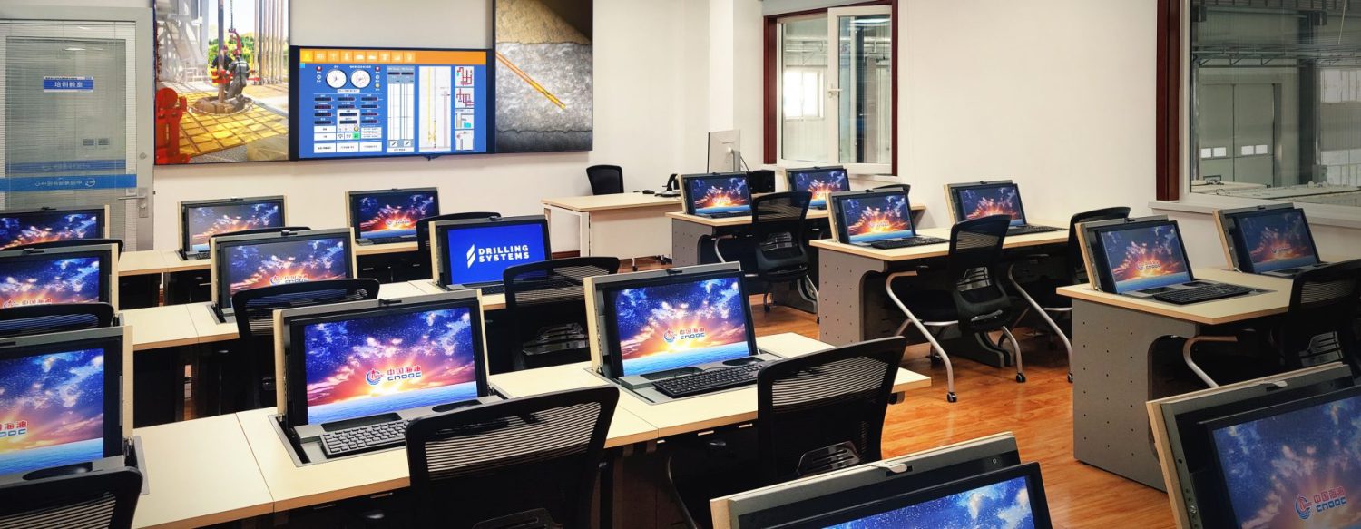 A computer lab with multiple rows of desks, each equipped with a desktop computer featuring brightly colored screensavers. The room includes a large display screen at the front.