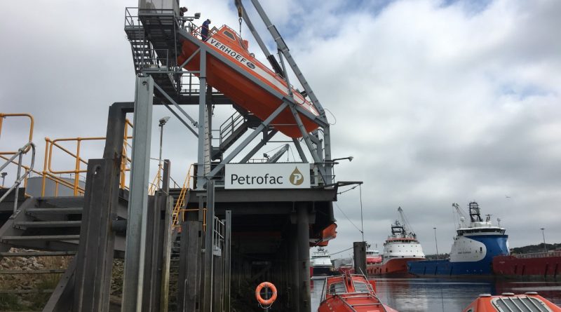 An orange lifeboat is suspended on a launching mechanism above the water at a Petrofac facility, with two ships docked in the background.