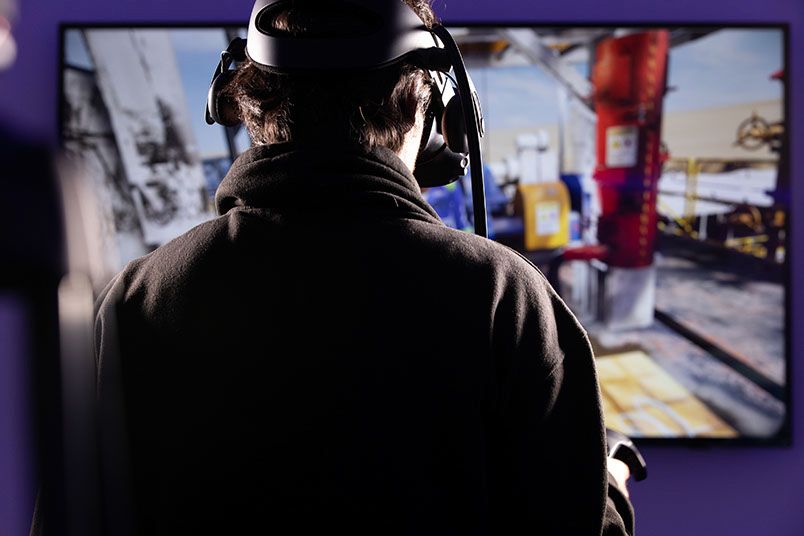 Person wearing a VR headset and holding controllers, engaging with a virtual reality experience. A screen in the background displays a colorful and dynamic scene.