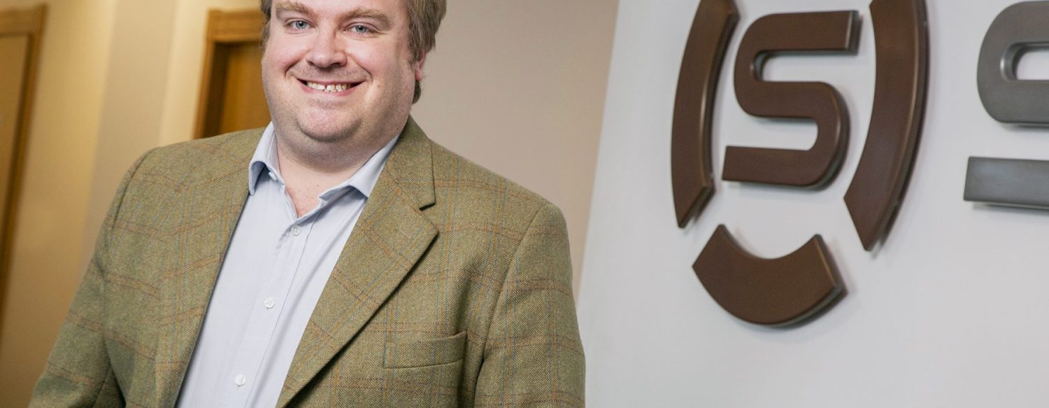 A man in a plaid blazer and light blue shirt stands smiling next to a wall with a partial logo featuring a stylized 'S'.