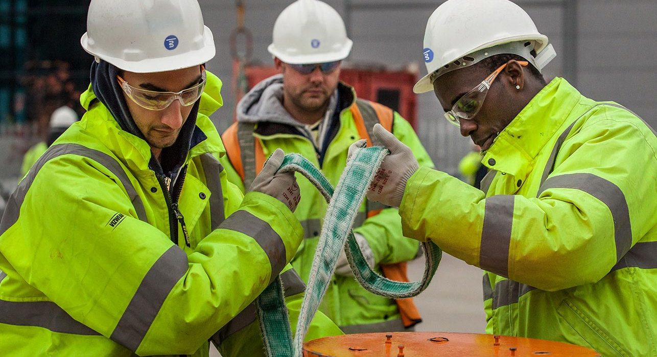 Three construction workers wearing hard hats and high-visibility jackets are securing a strap around an object at a construction site.