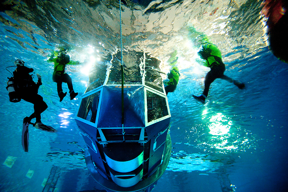 Several divers in scuba gear surround a submerged, large, windowed structure underwater, illuminated by bright lights.