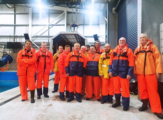 A group of people wearing orange and yellow safety attire stand indoors near a training pool facility.