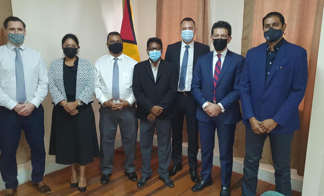 A group of seven people, wearing formal attire and face masks, stand in a room with curtains and a flag in the background.