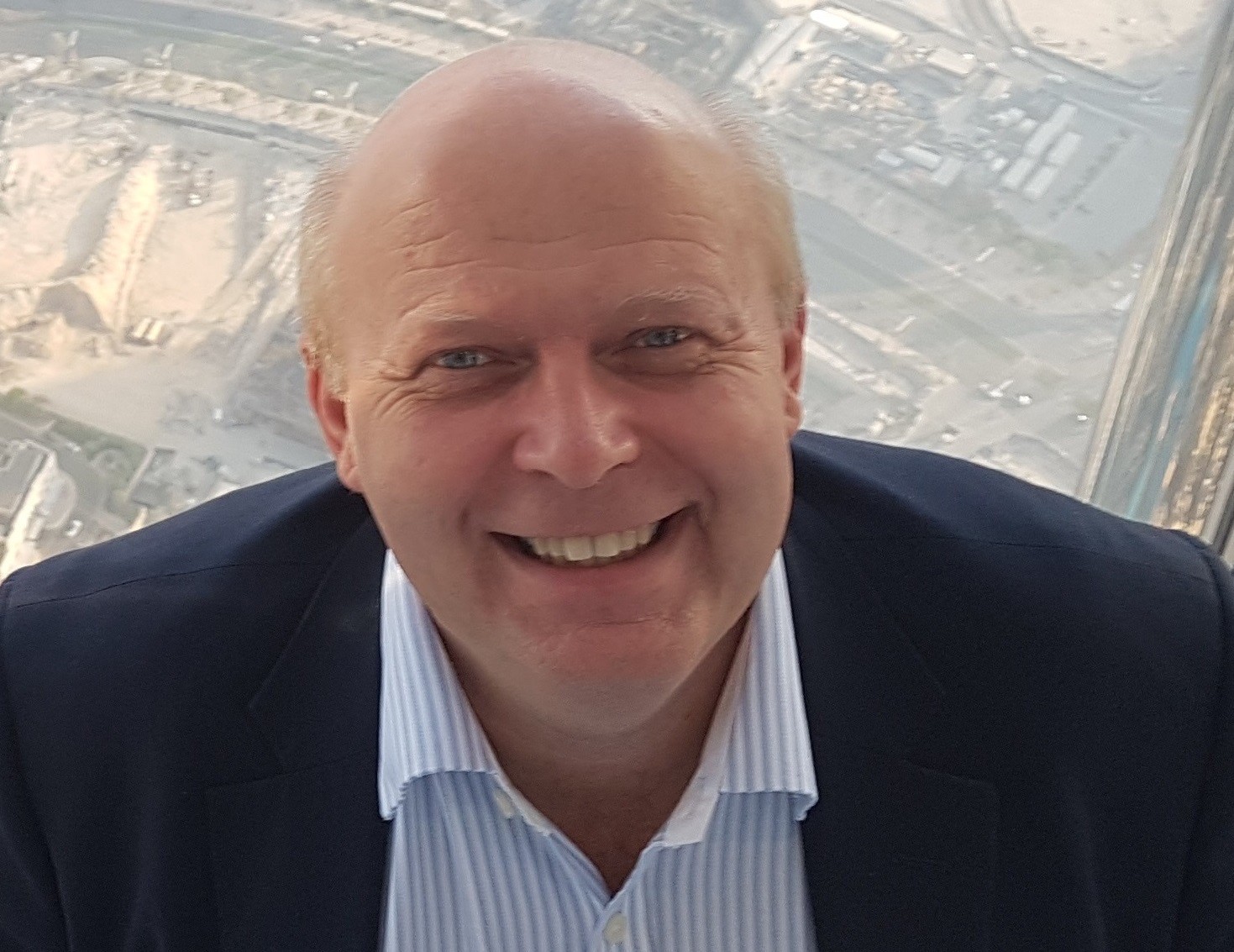 A smiling bald man in a dark suit and striped shirt is photographed from the chest up, with an aerial view of a city landscape in the background.