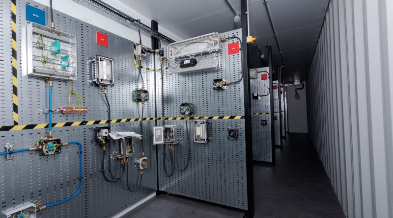 Industrial control room with various electrical panels mounted on perforated metal walls, featuring wiring, connectors, and caution tape. The room has a narrow walkway and industrial lighting.