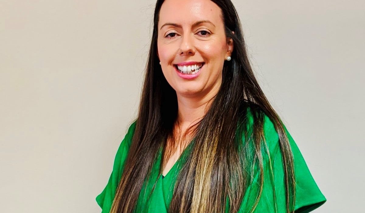 A smiling woman with long dark hair wearing a green blouse stands against a plain background.