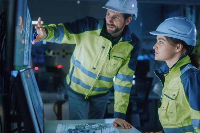 Two engineers wearing hard hats examine data on computer screens in a control room, with one pointing at the screen and the other attentively observing.