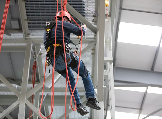 A person wearing safety gear and a red helmet is suspended by ropes, climbing on a metal structure inside an industrial facility.