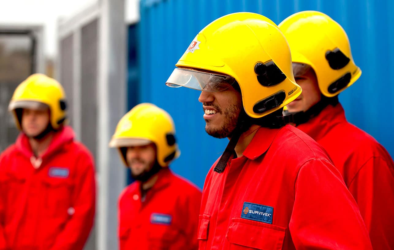 A group of four people wearing yellow helmets and red uniforms are standing outside beside blue structures.