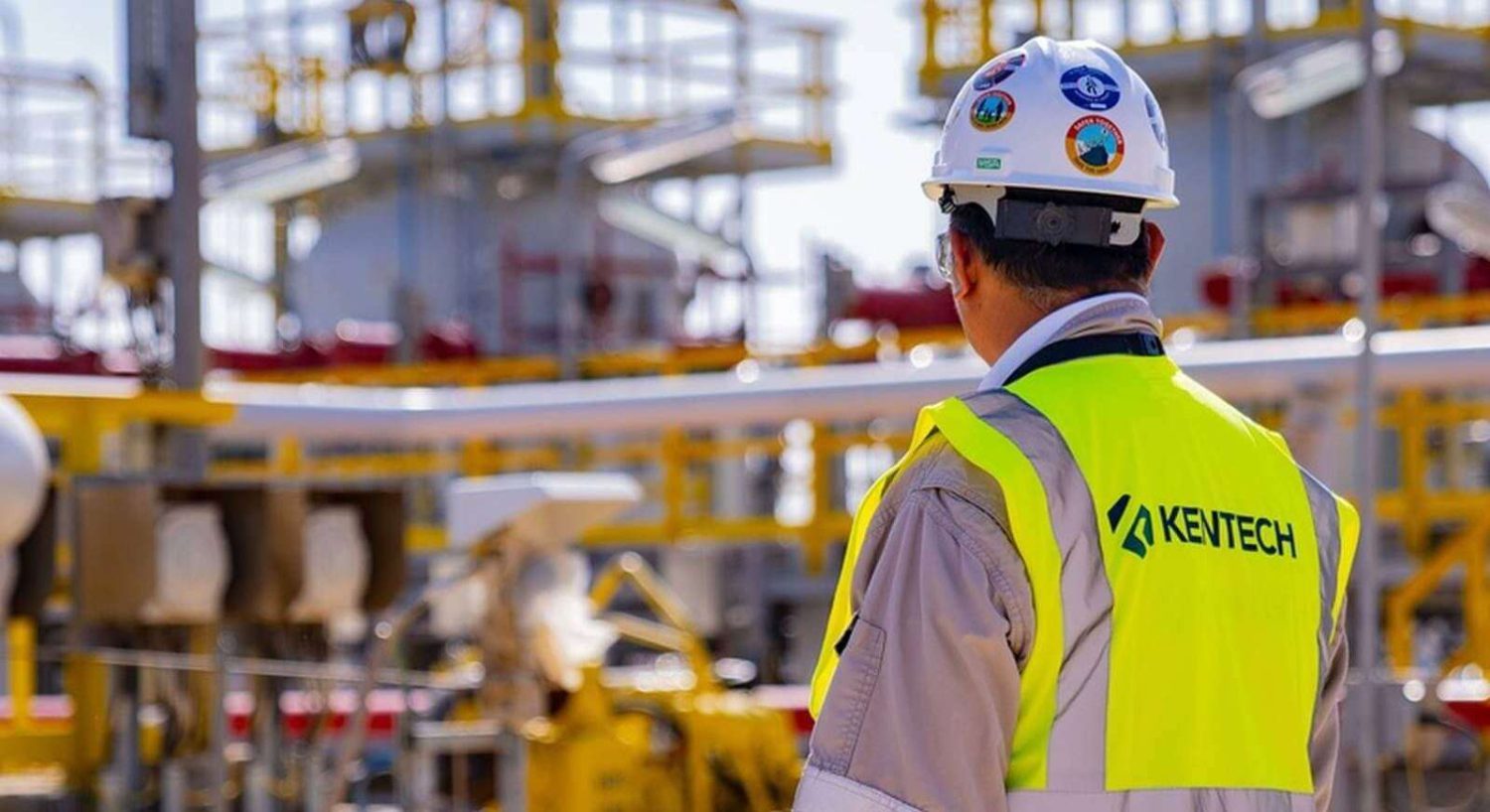 A worker wearing a high-visibility vest and a hard hat stands looking at an industrial site with various pipes and structures. The vest reads 