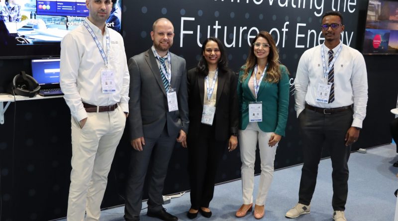 A group of five people, three men and two women, stand together for a photo at a business event. They are in front of a booth with displays and the text 