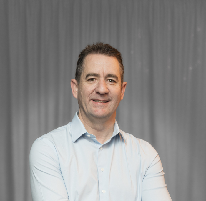 A man in a light blue shirt stands with his arms crossed, smiling, in front of a grey curtain background.