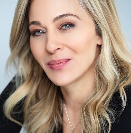 A woman with long, wavy blond hair and fair skin, wearing a black top, smiles softly.