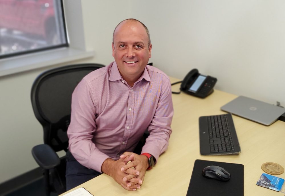 A man in a pink shirt sits at a desk with a computer keyboard, mouse, closed laptop, phone, pad of paper, and a drink coaster. He is smiling and looking at the camera.