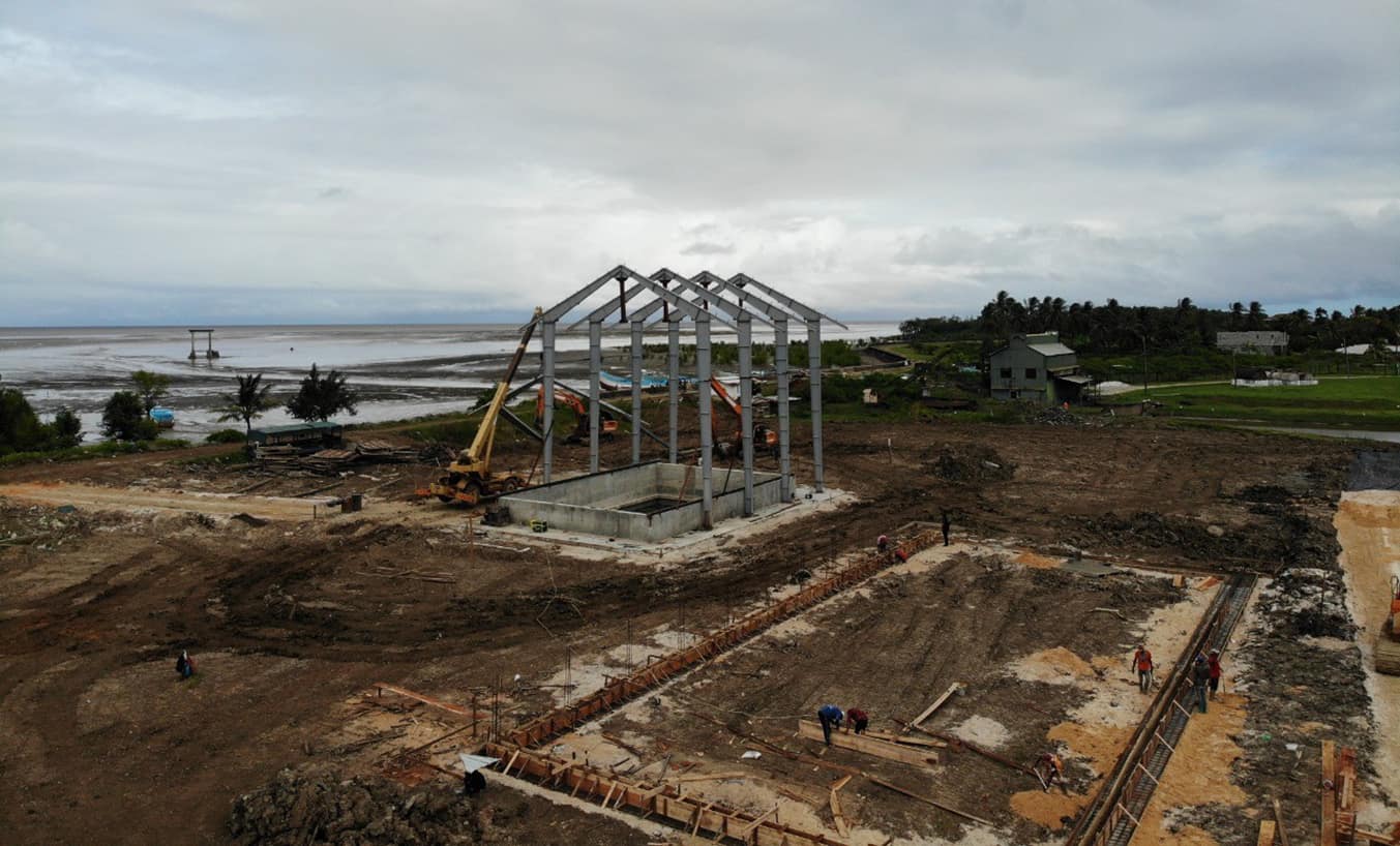 Aerial view of a construction site with steel framework in the center, surrounded by workers and machinery. The site is near a body of water and a small green building.