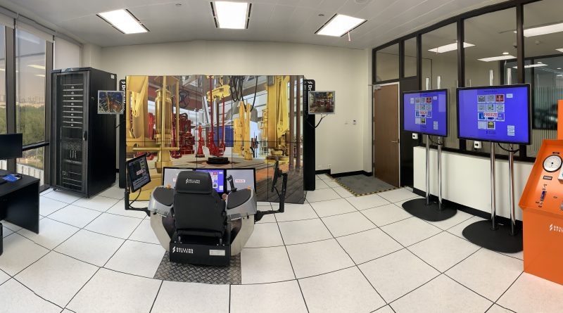 A room with a simulator setup, featuring a central control chair, multiple screens displaying various interfaces, and several other technical equipment and monitors around the room.