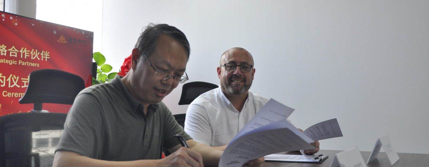 Two men are seated at a table with documents in front of them. One man is signing a paper, while the other smiles. A red banner with text in a foreign language is displayed in the background.