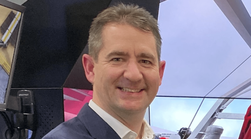 A man with short brown hair wearing a navy blazer and white shirt, smiling in front of a backdrop featuring modern design elements and technological equipment.