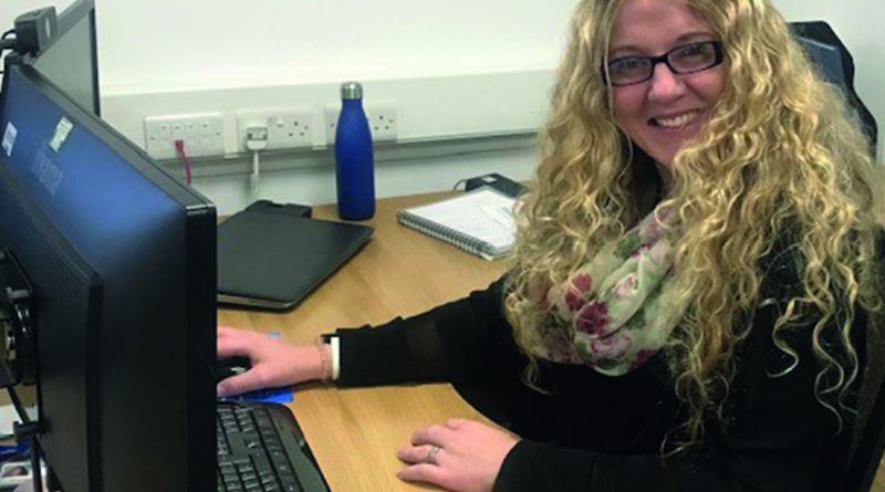 A person with long curly hair and glasses is sitting at a desk, smiling while using a computer. There is a blue water bottle, a notebook, and other office items on the desk.