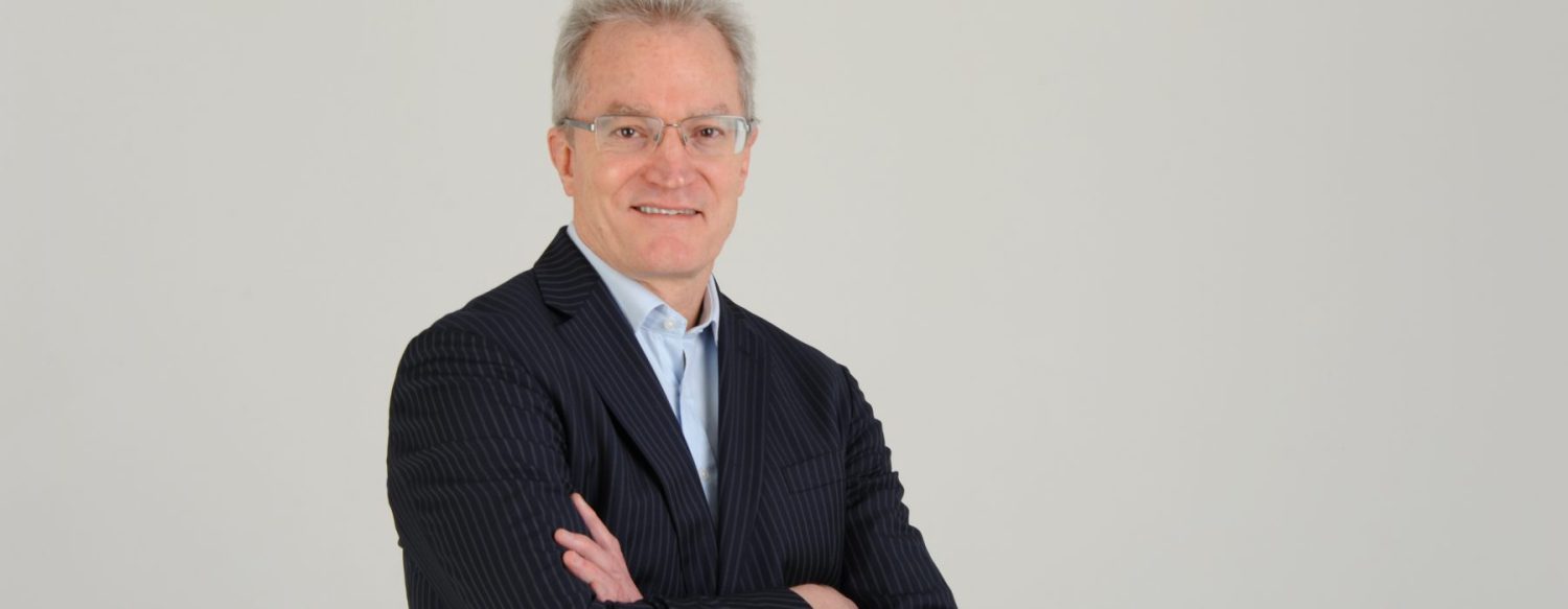 A man with short gray hair, wearing glasses, a light blue shirt, and a pin-striped dark suit, stands against a plain background with arms crossed, smiling at the camera.