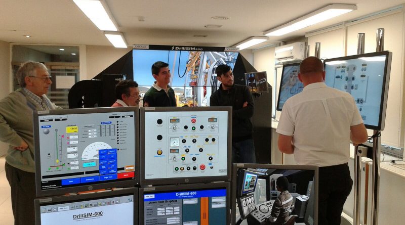 A group of people stand in a room with various monitors and control panels, attentively listening to a man giving a presentation in front of a large screen.