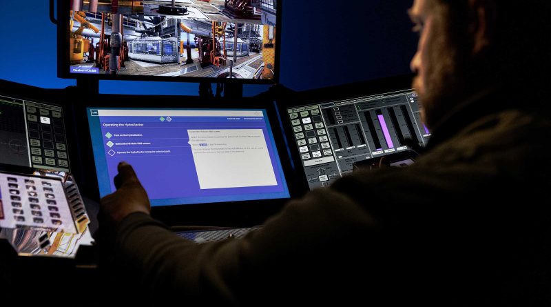 A person operates machinery using multiple computer screens and control panels in a control room, with industrial equipment visible on the monitors.