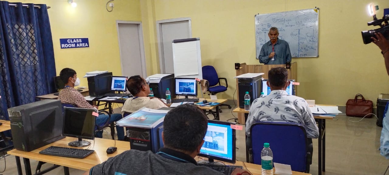 A man stands at a podium, speaking to a classroom of seated adults. The room has computer stations, a whiteboard with notes, and a sign reading 