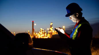 A worker in a hard hat and safety vest uses a tablet in a dimly lit industrial setting with bright lights in the background.