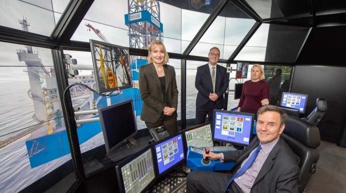 Four people are in a high-tech control room with several monitors displaying offshore drilling equipment. One person is seated, operating the controls, while the others stand behind, observing.