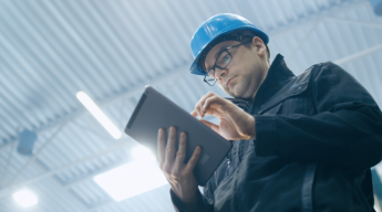 A person wearing a hard hat and glasses uses a tablet in an industrial setting with a corrugated metal ceiling.