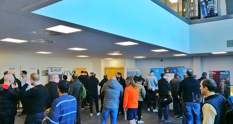 A group of people, some in conversation, stand in a spacious, well-lit lobby area with various informational banners and posters displayed around.