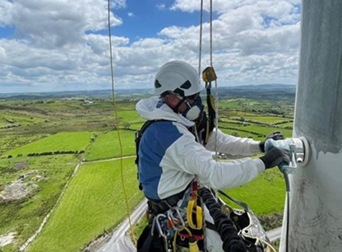 A person wearing safety gear and a helmet works on a structure high above a rural landscape with green fields under a partly cloudy sky.