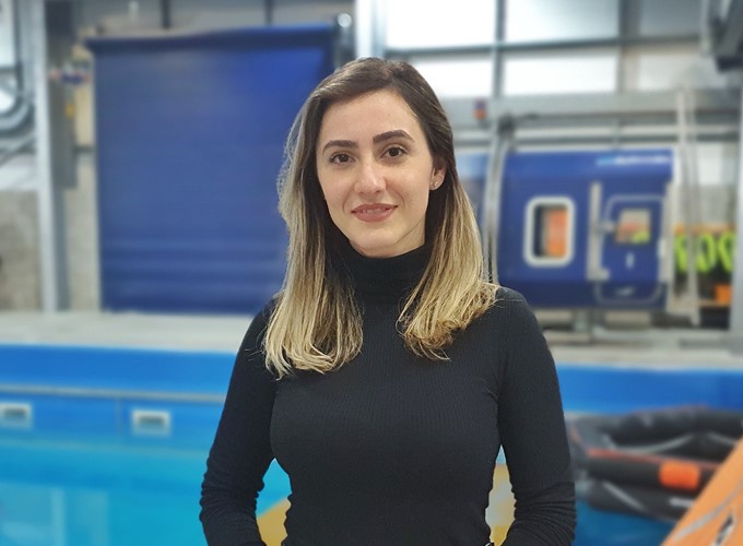 A person with long, light brown hair wearing a black turtleneck stands in front of a pool and an industrial blue background.