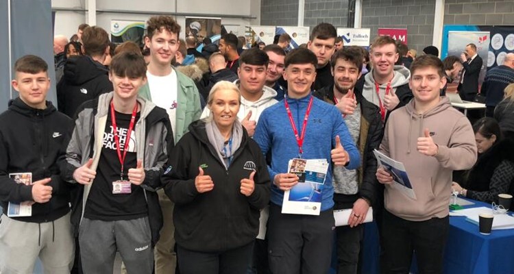 A group of people, mostly young men with a woman in the center, give thumbs up at an indoor event with informational booths and crowds in the background. They have event badges and brochures.