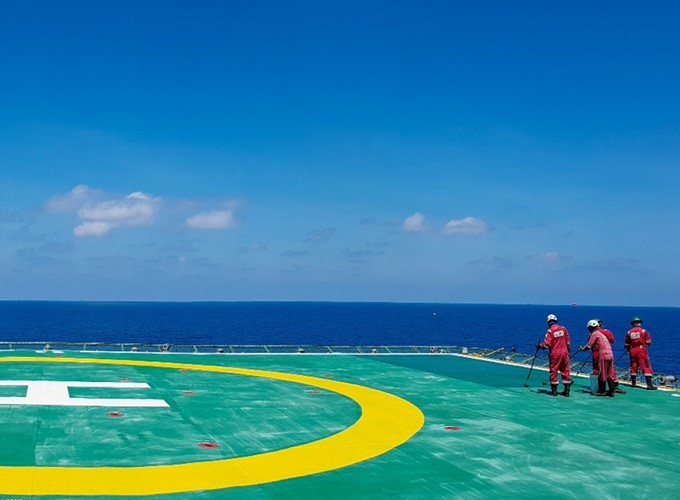 Three workers in red uniforms stand on a large helipad near the ocean under a clear blue sky.