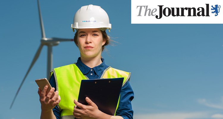 A person in a hard hat and high-visibility vest stands with a clipboard and phone, with a wind turbine in the background. 