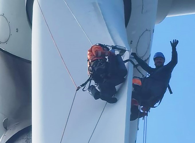 Two workers in safety gear are suspended on the blades of a wind turbine, with one worker waving at the camera.