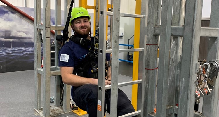 A man wearing safety gear and a helmet sits in a harness inside a metal structure indoors, with wind turbine models visible in the background.