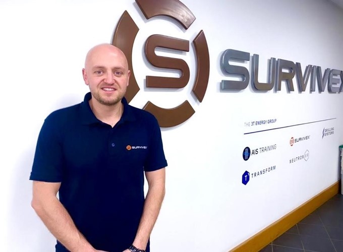 A person stands in front of a wall with the Survive logo and branding, dressed in a dark blue polo shirt with the same logo.