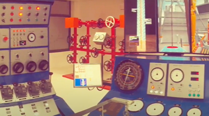 A control room with various dials, gauges, and equipment panels, situated behind a valve system mounted on a red frame.