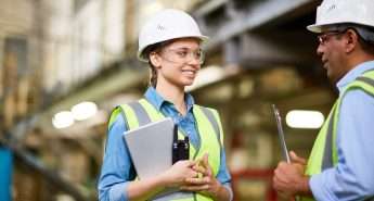 Two workers wearing hard hats and high-visibility vests have a conversation in an industrial setting. One holds a clipboard, and the other holds a wrench.