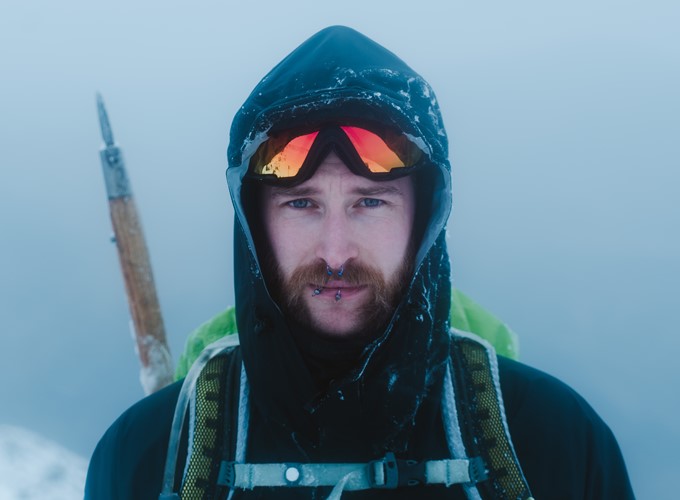 A person with facial piercings, sunglasses, and ice-covered clothing stands outdoors in snowy, foggy conditions. A hiking pole is attached to their backpack.