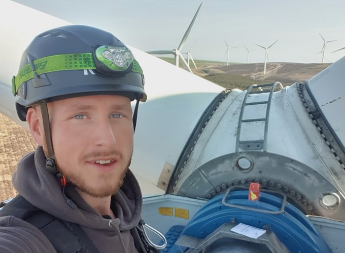 A person wearing a helmet and safety gear stands in front of a wind turbine, with multiple wind turbines visible in the background.