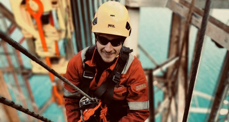 A worker wearing a yellow helmet and safety equipment is climbing a structure with ropes, with a large body of water visible in the background.