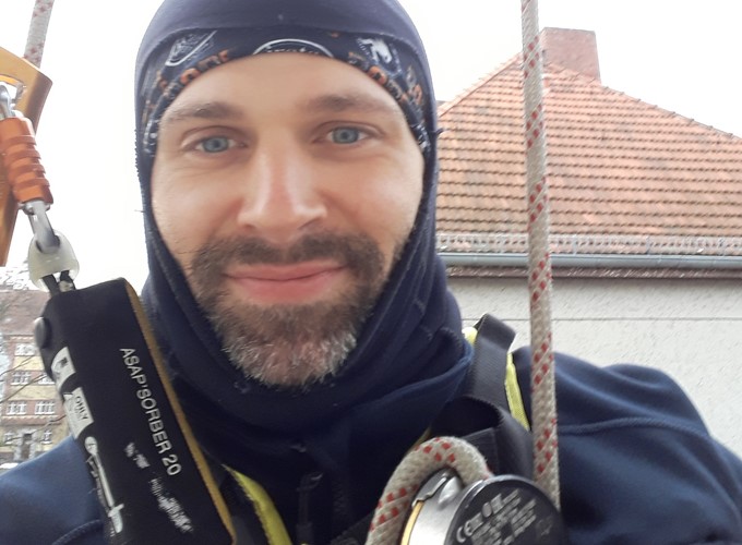 A person in outdoor gear, including a harness and ropes, smiles at the camera with a building and rooftop in the background.