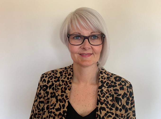 A person with short, light hair and glasses wearing a leopard-print jacket and a black top, standing against a plain background.