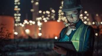 A worker wearing a hard hat and reflective vest uses a tablet in an industrial area at night. Illuminated structures in the background.